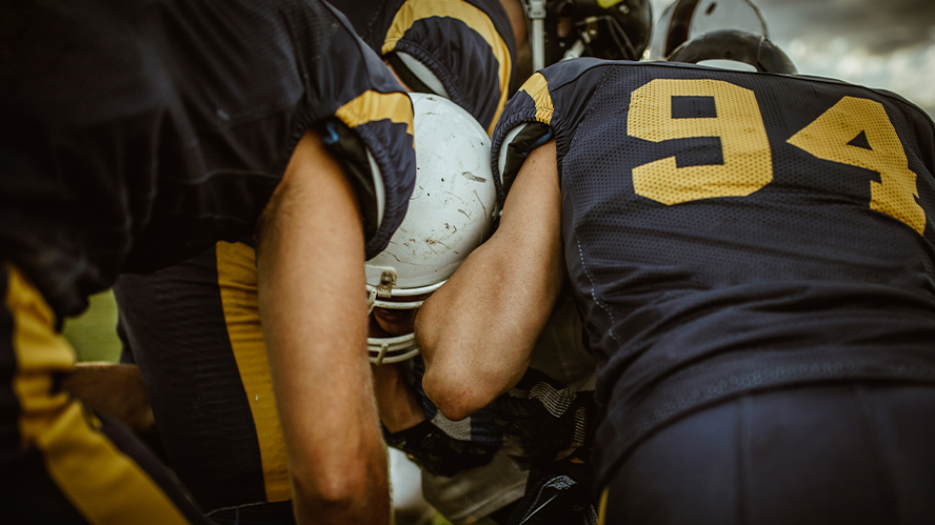 Close up image of American football pile up, with players in blue and yellow jerseys, and a white helemt featured in the centre